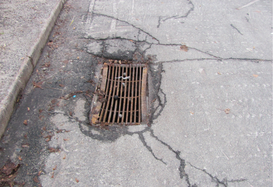 Rusted metal storm drain catch basin surrounded by broken asphalt and cracking pavement on a roadway.